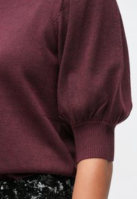 Μπορντό πλεχτό πουλόβερ με φουσκωτά κοντά μανίκια και ριμπ μανσέτες, φορεμένο με μαύρη φούστα από παγιέτες.