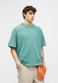 Νέος άνδρας με σγουρά μαλλιά, φορώντας μια φαρδιά τιρκουάζ μπλούζα και μπεζ σορτς, κρατώντας μια πορτοκαλί καπέλο στο αριστερό του χέρι.