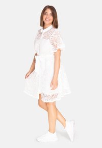Abito bianco in pizzo con colletto, maniche corte a campana, cintura legata in vita e orlo scallop. Indossato con sneakers bianche. Motivo floreale dettagliato su tutta la superficie.