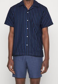 Camicetta a maniche corte con uno sfondo blu navy e strisce verticali blu, con chiusura a bottoni e taschino sul petto. Abbinata a pantaloni corti blu.
