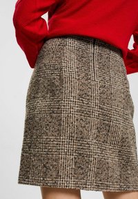 Hnědá kostkovaná sukně vyrobená z texturované látky, s rovný střih a lemováním těsně nad koleny, v kombinaci s červeným svetrem.