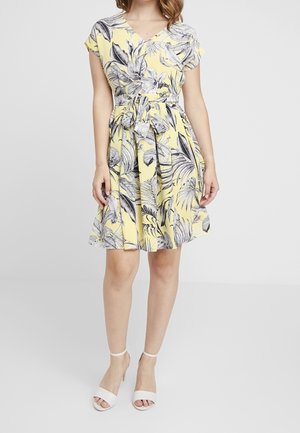 Žena v žlutých šatech po kolena s černobílými tropickými listy a látkovým páskem uvázaným v pase.