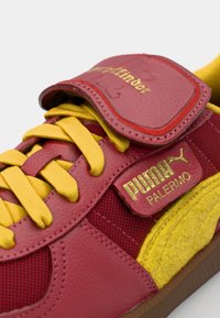 Sarkani un dzelteni Puma sporta apavi, izgatavoti no ādas un sieta materiāliem. Iezīmējas ar dzeltenām auklām, mīkstu sarkanu mēli un gumijas zoli ar zīmolu.