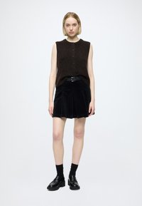 Hnedý bezrukávový sveter s gombíkmi, čierna plisovaná semišová sukňa, čierny opasok a mokasíny. Súčasťou sú čierne pančuchy po kolená.