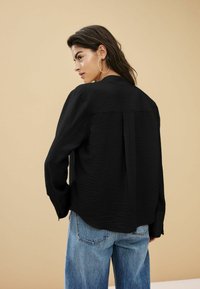 Sort tekstureret skjorte med løs pasform, knaplukning ved ærmerne og rygplissé, parret med lyseblå vide jeans.
