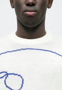 Witte gebreide trui met een geribde ronde hals, voorzien van een blauw abstract ontwerp in de stof geweven. De textuur oogt zacht en glad.