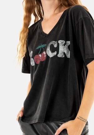 Osoba nosząca luźną czarną koszulkę V-neck z grafiką wiśni zastępującą literę "O" w słowie "ROCK."