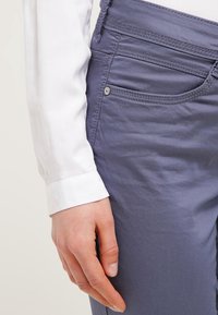 Jasnofioletowe spodnie slim-fit o gładkiej fakturze, z przednimi kieszeniami, detalami szycia oraz akcentem w postaci srebrnego guzika, zestawione z białą koszulą z długimi rękawami.