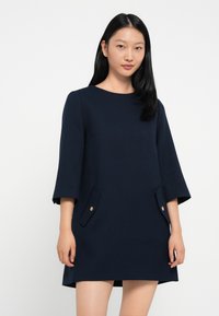Mørkeblå A-line kjole med trekvart ermer, teksturert stoff og to frontlommer pyntet med gullknapper.