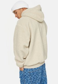 Felpa in pile di colore crema con una morbida e pelosa texture, caratterizzata da un cappuccio e polsini a coste, abbinata a jeans blu con motivo.