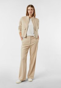 Beżowa kurtka bomberka na zamek z dwoma przednimi kieszeniami, w zestawie z beżowymi szerokimi spodniami. Biały T-shirt i białe trampki dopełniają strój.