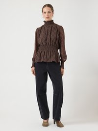 Blouse à manches longues à carreaux marron avec une taille froncée et un col haut à volants, associée à un pantalon noir à jambes larges et des talons beige.