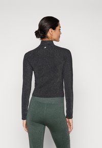 Sort langærmet top med høj krave og fin glimmertekstur, kombineret med grønne leggings. Subtilt logo på ryggen ved nakken.