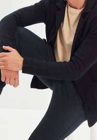 Cárdigan de punto azul marino con patrón texturizado, usado sobre una camiseta beige claro. Modelo sentado con pantalones negros, concentrado en una pose relajada.