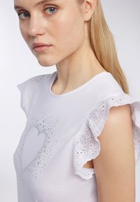 Top bianco con un design aderente, maniche a volant in pizzo e un dettaglio di strass a forma di cuore al centro, su uno sfondo chiaro.