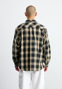 Camisa de franela con un patrón de cuadros en negro y beige, con mangas largas, cuello y detalles de botones, combinada con pantalones cargo blancos.