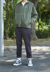 Lätt vikbar jacka i olivgrön med vita detaljer, kombinerad med mörka avsmalnande byxor och mönstrade grå och vita sneakers.