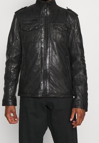 Zwarte leren jas met een hoge kraag, ritssluiting aan de voorkant, twee borstzakken en schouderaccenten. Gestructureerd oppervlak met een aansluitend ontwerp.