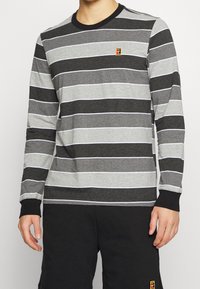 Graues Langarmshirt mit horizontalen schwarzen und hellgrauen Streifen, ausgestattet mit einem runden schwarzen Kragen und einem kleinen gestickten Logo auf der Brust.