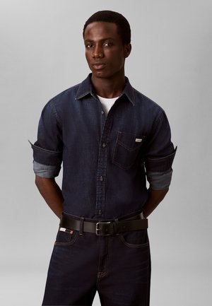 Unge mand med mørk hud og kort hår iført en mørk denimskjorte med op-rullede ærmer og mørke jeans, stående med hænderne bag ryggen.