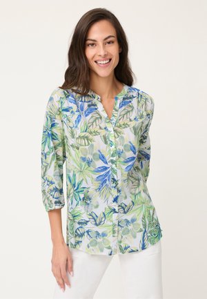 Lächelnde Frau trägt ein langärmeliges Hemd mit blau-grünem tropischem Blatt- und Blumenmuster, kombiniert mit weißen Hosen.