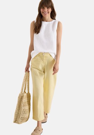 Smilende kvinde iført hvid ærmeløs top, gule vidde bukser, beige slip-on sko, der holder en stor vævet taske.