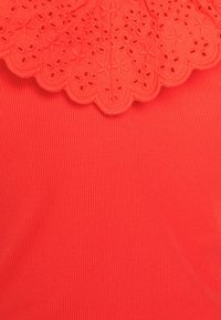 Röd topp med en kantad spetskrage med blommig broderi. Huvudtyget har en ribbad textur som förstärker den övergripande designen.