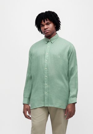 Jeune homme aux cheveux bouclés portant une chemise vert clair à boutons et un pantalon beige, debout devant un fond clair uni.