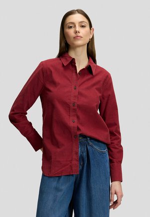 Frau steht mit der Hand in der Hüfte, trägt ein rotes Hemd mit Knöpfen und blaue hochgeschnittene Jeans vor einem schlichten hellgrauen Hintergrund.