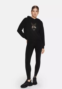 Black hoodie with a gold heart and bunny design, paired with black leggings and black sneakers, featuring a relaxed fit and hood.