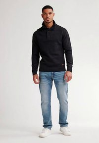 Svart stickad tröja med en knappad henley-krage och ribbstickade muddar, kombinerad med ljusblå jeans och vita sneakers. Neutral bakgrund.