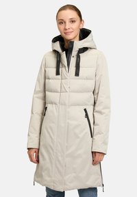 Manteau rembourré beige clair avec une capuche structurée, des accessoires noirs, deux zips latéraux et un motif matelassé sur la partie supérieure.