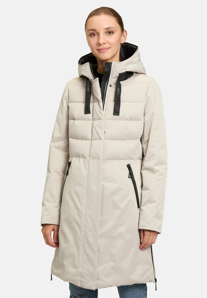 Manteau rembourré beige clair avec une capuche structurée, des accessoires noirs, deux zips latéraux et un motif matelassé sur la partie supérieure.