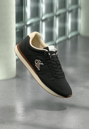 Black leather sneaker with tan accents and cream sole. Features a textured surface, lace-up front, and embroidered logo detail.