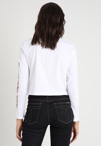 Weißes, kurz geschnittenes Langarmshirt mit Text auf dem linken Ärmel, kombiniert mit enganliegenden dunklen Jeans. Glattes Material, lässiges Design.