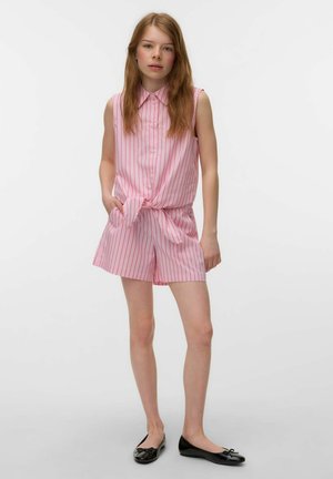 Młoda kobieta stojąca, ubrana w różowo-biały, pasiasty, bezrękawny top zapinany na guziki, przewiązany w talii oraz pasujące krótkie spodenki i czarne baleriny.