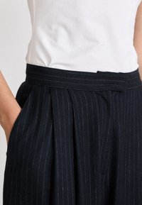 Pantaloni a righe navy con cintura ampia e pieghe, dotati di tasche laterali e una texture morbida. Una parte superiore bianca è parzialmente visibile.