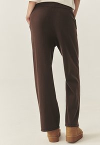 Bruine, lichte, taps toelopende broek met zakken, elastische tailleband en rechte pijpen. Gecombineerd met tan enkelbootjes op een lichte achtergrond.