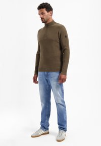 Gestreepte olijfgroene pullover met een halflange ritskraag, gecombineerd met lichtblauwe jeans en grijze sneakers met drie witte strepen.