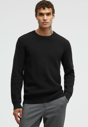 Schwarzer gerippter Strickpullover mit rundem Ausschnitt, langen Ärmeln und figurbetonter Silhouette, getragen zu grauen karierten Hosen. Glatte Textur.