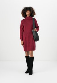 Frau mit lockigem Haar, die ein rotes Strick-Turtleneck-Kleid, schwarze knielange Stiefel trägt und eine schwarze Schultertasche hält, steht vor einem weißen Hintergrund.