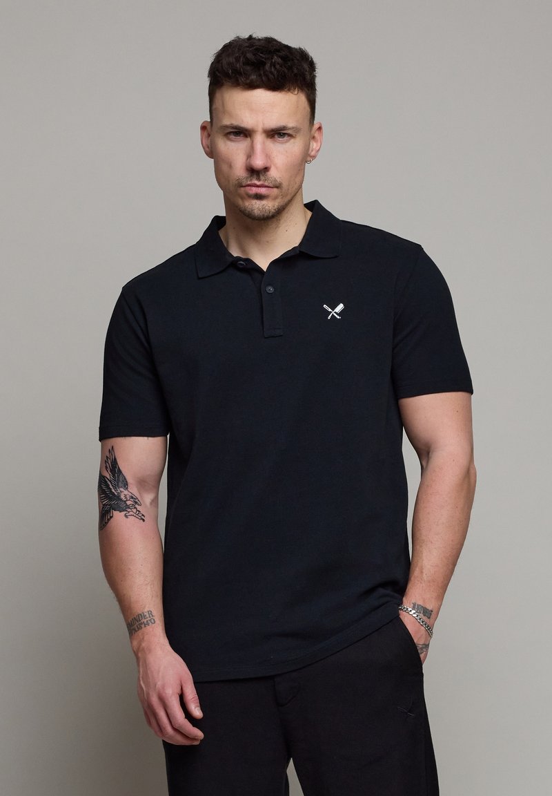 Homme aux cheveux courts portant un polo noir avec un logo de batte et balle croisées, montrant des tatouages sur les deux avant-bras, fond neutre.