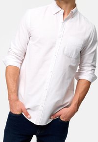 Chemise blanche à boutons en coton avec une poche sur la poitrine gauche, manches longues retroussées au niveau des poignets. Portée avec un jean bleu foncé.