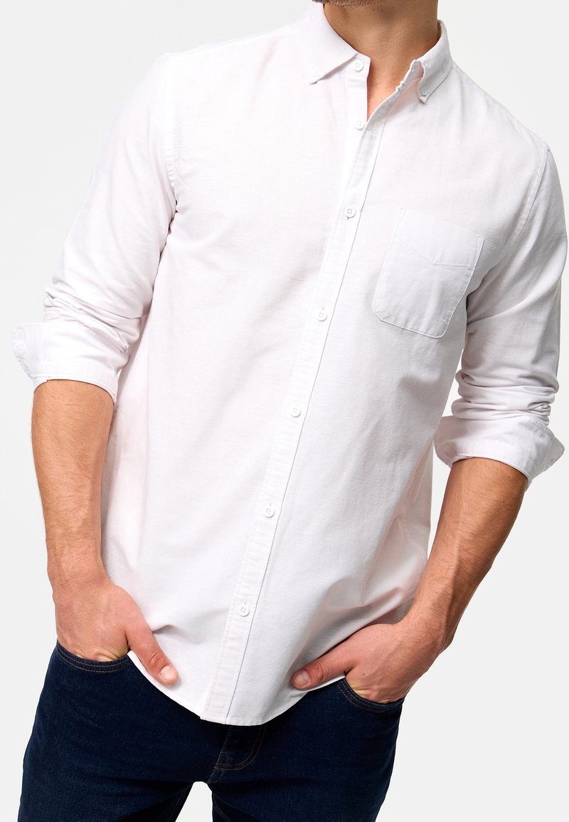Chemise blanche à boutons en coton avec une poche sur la poitrine gauche, manches longues retroussées au niveau des poignets. Portée avec un jean bleu foncé.