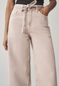 Világos rózsaszín farmerből készült, bő szárú nadrág. Magasított derékrésszel, megkötő részlettel, oldalsó zsebekkel és finoman kopott hatású textúrával rendelkezik.