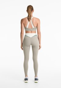 Vrouw met een staart die een beige sportbeha met racerback en hoge taille leggings draagt, staande met haar rug naar de camera op een witte achtergrond.