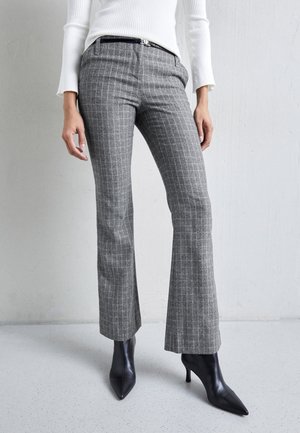Kobieta w szarych, kraciastych dopasowanych spodniach z rozszerzanymi nogawkami, czarnych czółenkach na obcasie z ostrym noskiem oraz białym, prążkowanym longsleeve.