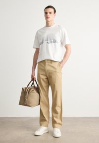 Hvit t-skjorte med dyretrykk, beige bomullsbukser, hvite sneakers og en brun veske med stropper. Enkel design og nøytrale farger.