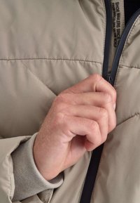 Chaqueta acolchada beige con diseño de acolchado. La mano está subiendo la cremallera frontal con una cremallera oscura. Un puño gris es visible debajo.