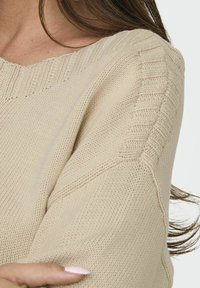Beige kötött pulóver széles nyakkivágással és bordás vállrész részletekkel. Puha textúra, laza szabás és varrás részletek az ujjakon.